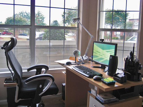 a desk with a computer in an office chair in front of a window